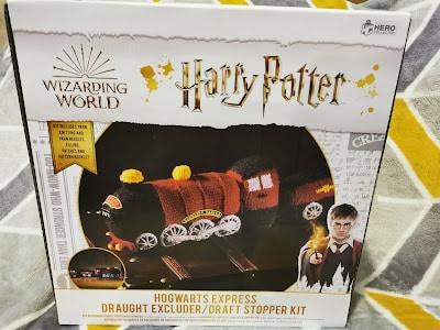 A boxed craft kit labelled "Wizarding World Harry Potter Hogwarts Express Draught Excluder/Draught Stopper Kit." The front shows a knitted Hogwarts Express train. Harry Potter, in robe and holding his wand, appears at the bottom right. Geometric patterned background.