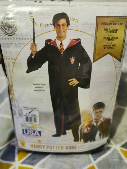 A packaged Harry Potter costume shows a smiling man wearing glasses, holding a wand, and dressed in a black robe with a red and gold Gryffindor emblem. Gold label reads “Hooded Robe with Clasp.” Harry Potter image and text "Harry Potter Robe" shown below.