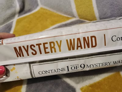 A hand with pink painted nails holds a rectangular package labelled "MYSTERY WAND." Text below reads "CONTAINS 1 OF 9 MYSTERY WANDS." The background is a soft blanket with a hexagonal yellow, grey, and white pattern.