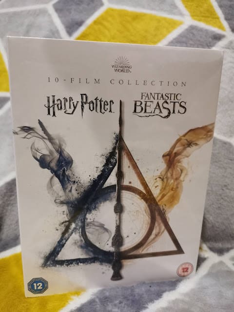 A DVD box set cover titled "10-Film Collection: Harry Potter Fantastic Beasts" features smoky blue and gold wisps forming magical symbols, with a wand forming a triangle and circle. The cover sits on a geometric-patterned fabric background. Age rating 12.