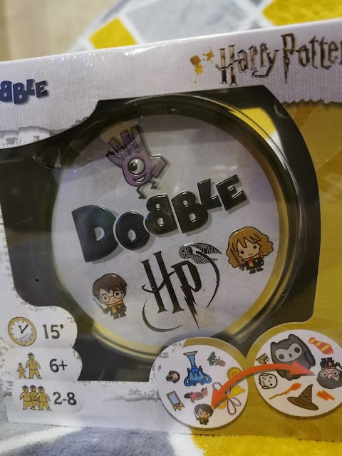 A boxed “Harry Potter Dobble” card game. The packaging shows the logo with cartoon icons of Harry, Hermione, and magical objects. Symbols denote game time (15 min), age (6+), and players (2-8). Part of the yellow patterned background is visible.