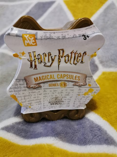 A Yume Harry Potter Magical Capsules Series 1 toy container sits on a textured yellow, grey, and white patterned surface. The packaging is shaped like a scroll and features gold lettering and accents, with the Wizarding World logo at the bottom.