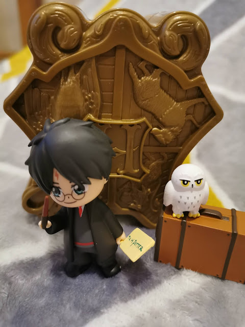 A Harry Potter figurine holds a wand and a letter, standing next to a small white owl atop a brown suitcase. Behind them is a brown Hogwarts crest with an “H” and engravings of the four house mascots. The background is a soft, patterned blanket.