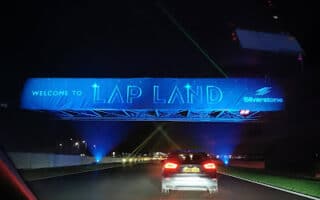A night scene shows a car driving under a blue-lit overhead sign that reads "Welcome to Lap Land" with the Silverstone logo. The road is illuminated and bordered by grassy strips, suggesting a festive event at a racetrack.