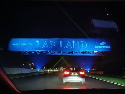 A night scene shows a car driving under a blue-lit overhead sign that reads "Welcome to Lap Land" with the Silverstone logo. The road is illuminated and bordered by grassy strips, suggesting a festive event at a racetrack.