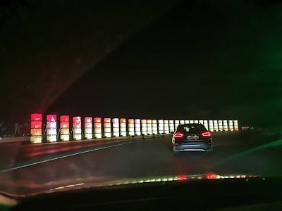 A night-time scene shows a car driving on a road. Ahead, a row of illuminated vertical panels in red, yellow, and green lines the roadside. The photo is taken from inside another car, with the dashboard faintly visible at the bottom.