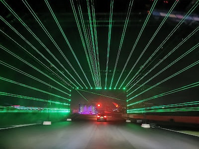 A car drives through a roadway tunnel at night, illuminated by bright green laser lights forming parallel beams overhead and to the sides. In the distance, colourful lights and more cars are visible, creating a futuristic, vibrant atmosphere.