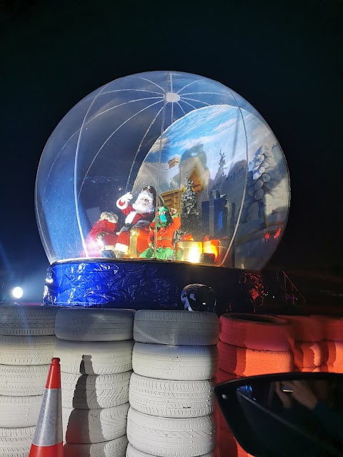 A large illuminated snow globe featuring Father Christmas and festive decorations sits atop stacked white and red tyres at night. An orange traffic cone is in the foreground, and a partial car window is visible at the bottom right.