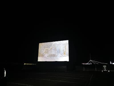 A large outdoor cinema screen displays a snowy winter scene with text at the bottom, set against a dark night sky. The screen is in an empty drive-in theatre with faint outlines of tents and parked cars visible in the darkness.