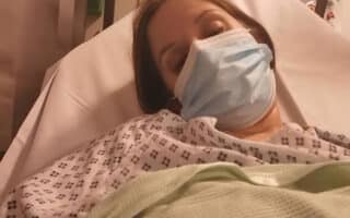 A person lies in a hospital bed, wearing a hospital gown and a blue surgical mask, partially covered with a green blanket. Medical equipment and a white wall are visible in the background. The person appears tired or resting.