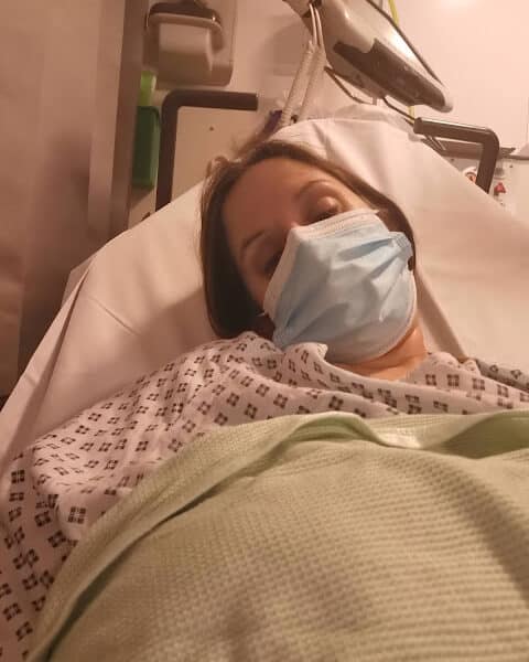 A person lies in a hospital bed, wearing a hospital gown and a blue surgical mask, partially covered with a green blanket. Medical equipment and a white wall are visible in the background. The person appears tired or resting.