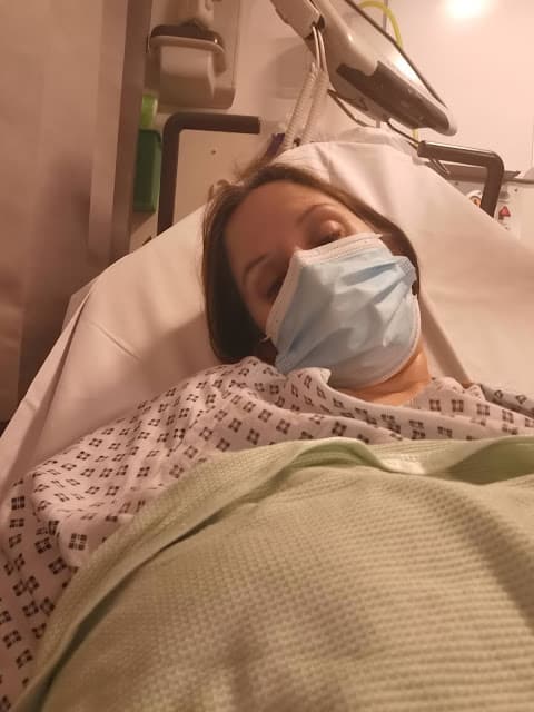A person lies in a hospital bed, wearing a hospital gown and a blue surgical mask, partially covered with a green blanket. Medical equipment and a white wall are visible in the background. The person appears tired or resting.