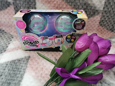 A boxed set of two colourful bath bomb kits labelled "So Bomb DIY" rests on a soft, patterned blanket. In front of the box is a small bouquet of artificial purple tulips tied with a purple ribbon.