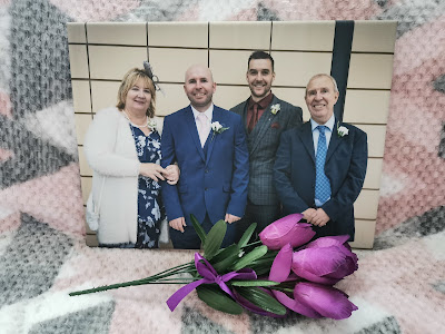 A family photo of two men in suits, an older man in a blue suit, and a woman in a white jacket, smiling in front of a tiled wall. The framed photo is placed on a pink, grey, and white blanket, with three purple tulips tied with ribbon in front.