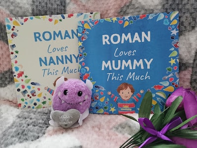Two colourful books with cartoon designs read “ROMAN Loves NANNY This Much” and “ROMAN Loves MUMMY This Much.” A plush purple toy holding a heart and a purple artificial tulip lie in front, all on a soft, chequered blanket background.