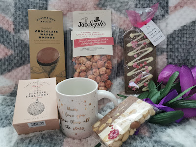 A gift assortment on a rug includes chocolate wafer rounds, caramel white chocolate popcorn, a chocolate bar, Earl Grey tea, a “Mum I love you” mug, a biscuit, and a bouquet of purple tulips, arranged together in a cosy setting.