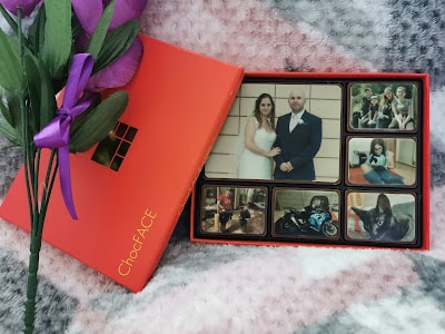 A box of ChocFACE chocolates featuring edible photo prints of people, a cat, and a motorbike sits open on a soft, patterned blanket. Purple artificial flowers with green leaves lie beside the bright red box.