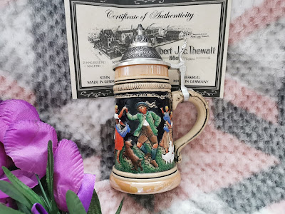 A decorative German beer stein with a pewter lid features a colourful scene of people dancing. Behind it, a Certificate of Authenticity is partially visible. In the foreground, artificial purple flowers rest on a checked blanket with pink, grey, and white tones.