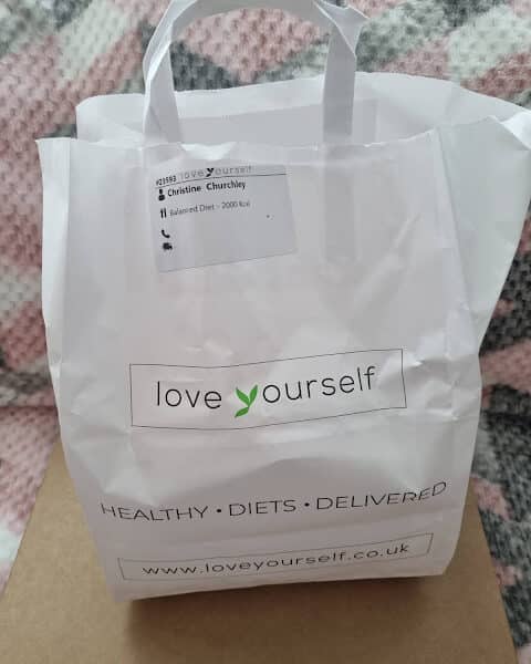 A white paper bag with handles sits on a beige surface, in front of a pink-and-grey checked blanket. The bag reads “love yourself” and “HEALTHY . DIETS . DELIVERED” with the website “www.loveyourself.co.uk.” A label with a name and diet information is attached.