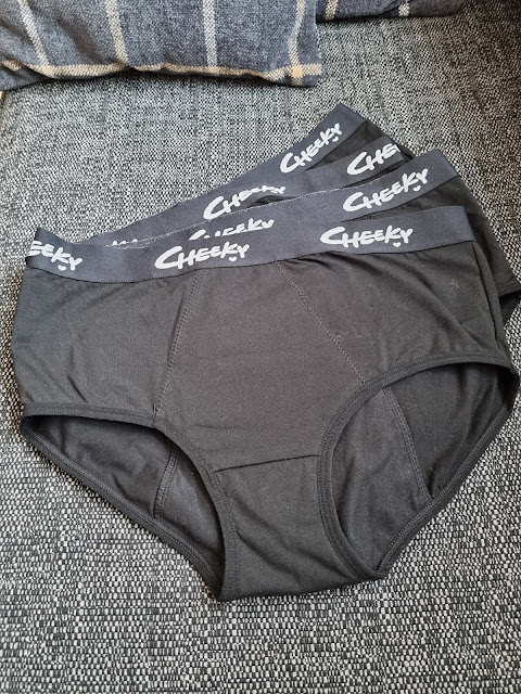 Two pairs of black underwear with “Cheeky” written in white on the waistband are stacked on a textured grey surface. A corner of a grey and white striped blanket is visible at the top of the image.