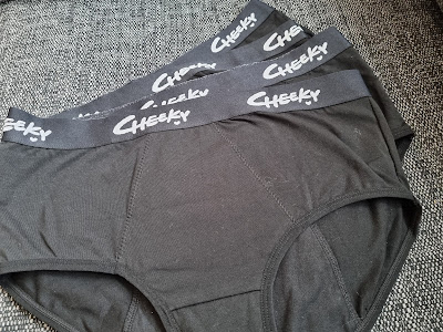 Three pairs of black knickers are stacked on a grey fabric surface. Each waistband is black with the word "Cheeky" printed in white, cursive-style letters. The knickers appear new and neatly arranged.