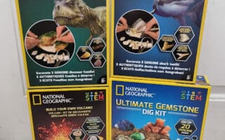 Four National Geographic STEM dig kits are shown: Dino Fossil, Shark Tooth, Build Your Own Volcano, and Ultimate Gemstone Dig Kit. Each colourful box displays related images, kit components, and educational age recommendations against a white background.