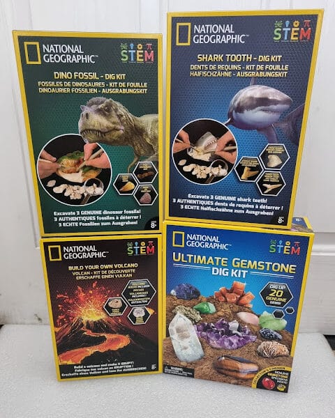 Four National Geographic STEM dig kits are shown: Dino Fossil, Shark Tooth, Build Your Own Volcano, and Ultimate Gemstone Dig Kit. Each colourful box displays related images, kit components, and educational age recommendations against a white background.