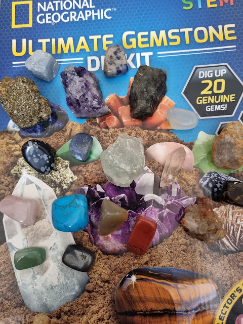 A variety of colourful gemstones are arranged on top of a "National Geographic Ultimate Gemstone Dig Kit" box. The box shows soil, crystals, and text about digging up 20 genuine gems, with stones of different shapes, colours, and sizes displayed.