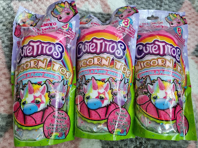 Three colourful Cutetitos Unicornitos toy packages stand upright on a soft, pastel-patterned surface. Each package features a cartoon unicorn face with a rainbow mane, pink cheeks, and blue nose, and promises to reveal a furry unicorn friend inside.