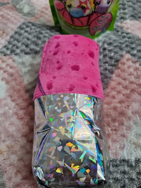 A shiny, holographic package wrapped partly in a pink, sponge-like cloth sits on a textured, chevron-patterned blanket. In the background, a colourful bag with cartoon animal faces is partially visible.