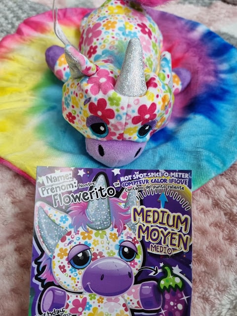 A colourful plush dragon with a silver unicorn horn, covered in multicoloured flower patterns, sits on a pastel tie-dye mat. In front, a card displays an illustration of the same dragon and the name "Flowerito," labelled as "Medium" spiciness.