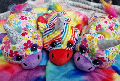 Three plush unicorn toys lie side by side on a tie-dye blanket. Two have pastel floral patterns, silver horns, purple snouts, and blue eyes. The middle unicorn features a rainbow-striped body, red snout, blue eyes, and a silver horn.