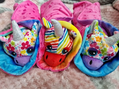 Three colourful plush unicorn toys with glittery silver horns and floral or rainbow-patterned faces lie side by side on a soft, pink, and grey blanket. Each unicorn is wrapped in a bright hooded blanket matching its unique design.
