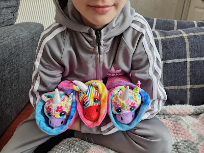 A child in a grey Adidas tracksuit sits cross-legged, holding three colourful plush unicorns with different patterns: one has rainbow stripes, one is tie-dyed, and one is covered in bright floral designs. The child’s face is half visible, smiling slightly.