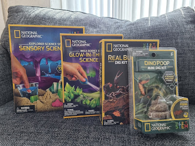 Four National Geographic science kits are displayed on a grey sofa: Sensory Science, Glow-in-the-Dark Science, Real Bug Dig Kit, and Dino Poop Mini Dig Kit. The boxes feature colourful, engaging graphics and are upright in a row.
