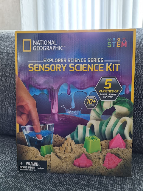 The image shows a box for the National Geographic Explorer Science Series Sensory Science Kit, featuring colourful sand, slime, and putty. The box displays a hand holding a clear cup, sand shapes, and lists “5 varieties of sands, slimes & putty” for ages 10+.