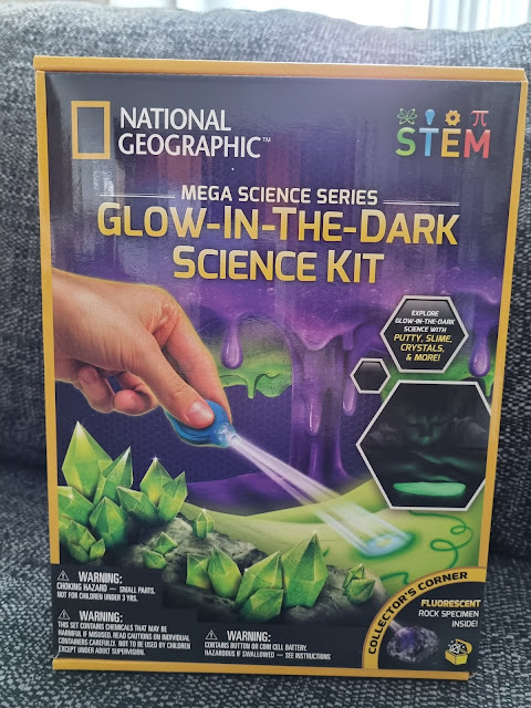 A box for the National Geographic Mega Science Series "Glow-in-the-Dark Science Kit." The cover shows a hand using a tool on green glowing crystals, with purple slime dripping in the background. The kit highlights STEM learning and includes a fluorescent rock specimen.