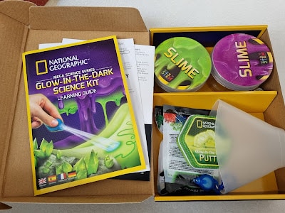 Open box containing a National Geographic Glow-in-the-Dark Science Kit. Inside are a learning guide, two containers labelled "slime," bags of materials, and a plastic funnel. The kit packaging is colourful with green and purple accents.