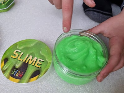 A person uses one finger to touch bright green slime in a clear plastic container. The container lid, labelled "SLIME" with a gooey green design and "STEM" logo, lies nearby on a white textured surface. Only hands and part of arms are visible.