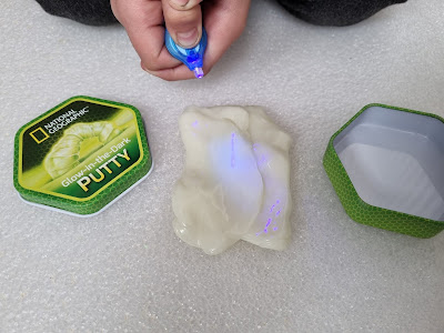 A person uses a small blue UV torch to draw on a mound of glow-in-the-dark putty. The putty glows faintly under the torch. Next to it is an open green National Geographic Putty tin on a white textured surface.
