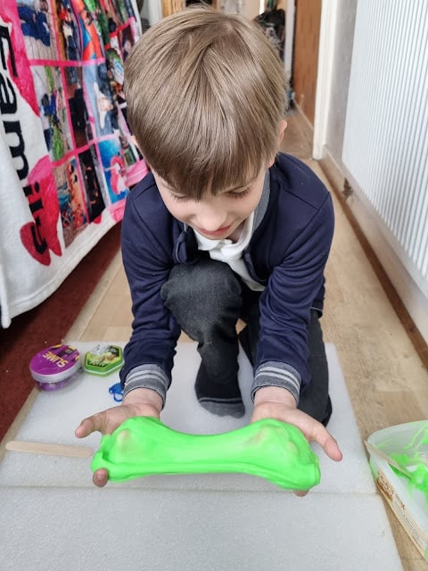 A young boy with light brown hair kneels on a white mat indoors, stretching bright green slime between his hands. He is wearing a navy blue cardigan and grey trousers. Colourful containers and toys are scattered nearby on the floor.
