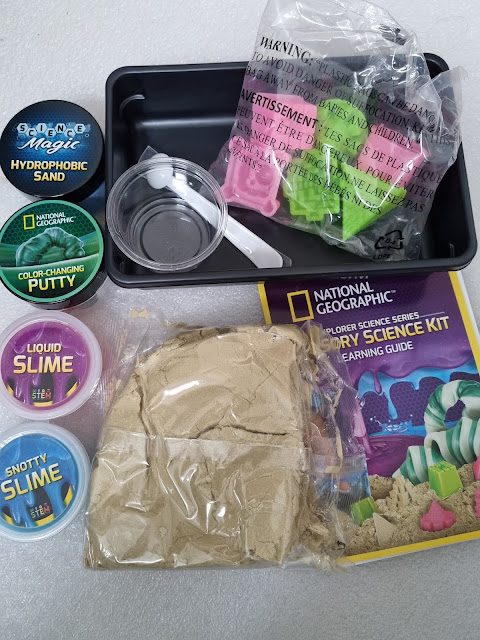 A National Geographic sensory science kit includes hydrophobic sand, colour-changing putty, two tubs of slime (liquid and snotty), small plastic moulds, a plastic tray, measuring spoons, and a colourful activity guide. All items are arranged on a white surface.