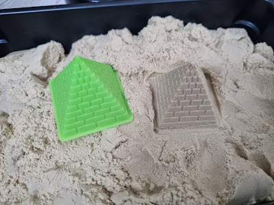 A green plastic pyramid mould sits on tan kinetic sand, next to a pyramid-shaped sand impression with a brick pattern, created by pressing the mould into the sand. Both objects rest in a black tray.