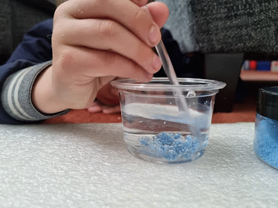 A child's hand stirs a clear plastic cup filled with water and blue crystals using a plastic stick. A small container of blue crystals sits nearby on a white textured surface, suggesting a science experiment or craft activity.