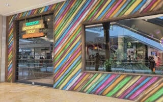 The entrance to Treetop Adventure Golf features a facade with colourful diagonal stripes, a large window displaying reflections, and a neon sign above the door. The scene is inside a shopping centre, with escalators and shopfronts visible in the background.