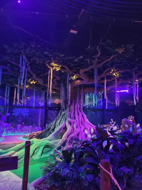 A large artificial tree with glowing purple and blue lighting stands in an indoor mini-golf area. The tree has thick roots, hanging vines, and is surrounded by green turf and leafy tropical plants, creating a mystical, fantasy-like atmosphere.