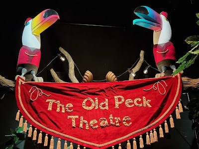 Two colourful toucan puppets wearing red jackets perch on branches above a red, tasselled banner that reads "The Old Peck Theatre" in yellow letters. The background is dark, with some green leaves at the edge and spotlights pointed at the scene.