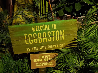 A wooden sign among lush green plants reads: "Welcome to EGGBASTON, twinned with Squawk City." Below it, a smaller sign says, "Stay put before you putt. Wait on your hole until the hole in front is free." The scene appears tropical and playful.