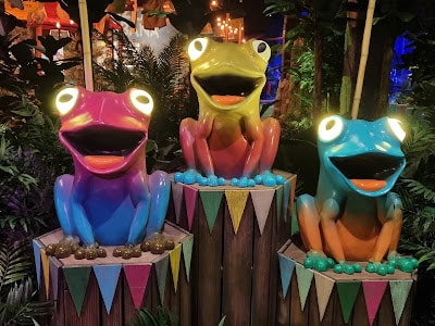 Three brightly coloured frog statues with glowing eyes sit on decorated wooden pedestals. Each frog is a different colour: pink and purple, green and yellow, and blue and orange. They are surrounded by jungle foliage and string lights in the background.