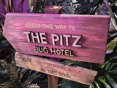 A wooden sign with purple tones reads, "Creep this way to The Pitz Bug Hotel." A smaller, weathered plank below says, "Shady rooms available in this dive hive," surrounded by green foliage and plants in the background.